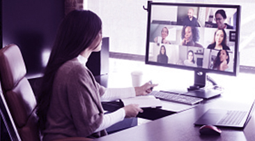 Nainen työpöydän ääressä. Tietokoneen ruudulla videopuhelu kahdeksan muun henkilön kanssa.